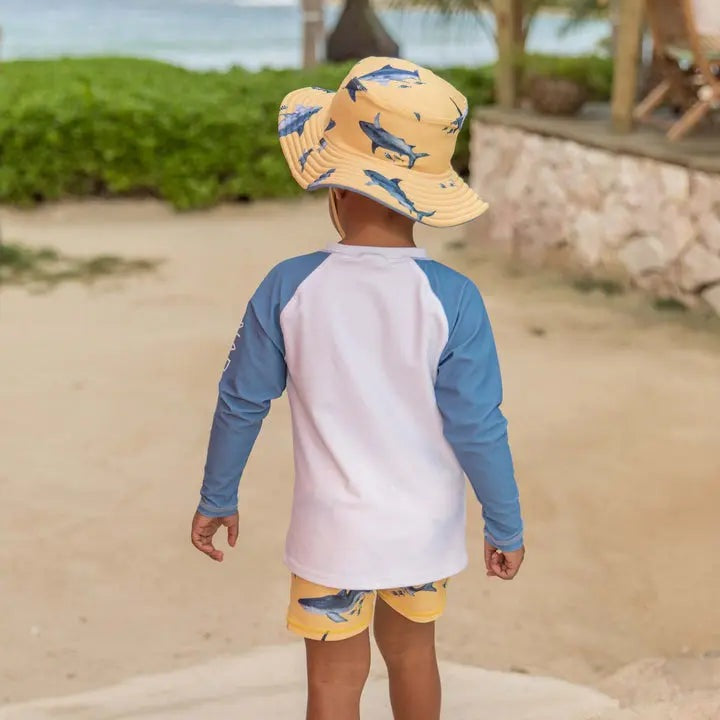 Snapper Rock Sunrise Shark Baby Set: toddler walking on the beach in blue raglan sleeves, white tee, and yellow shark-print shorts.