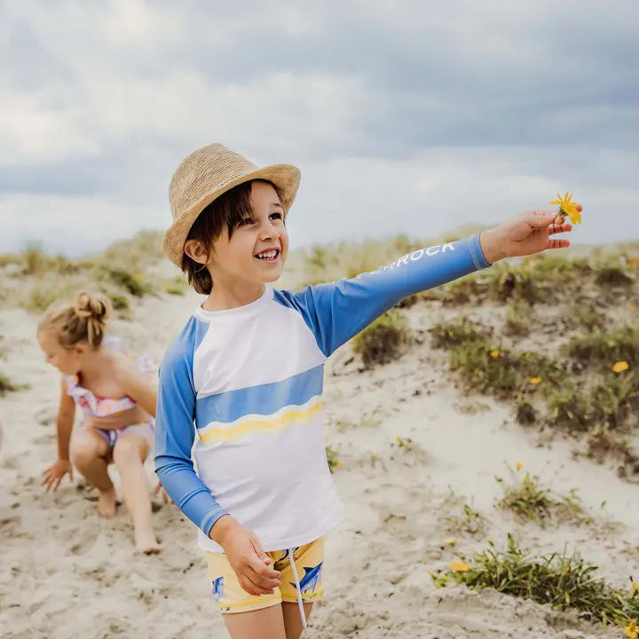 Snapper Rock Sunrise Shark Baby Set: child on a sunny beach in blue rash guard with yellow stripe and matching yellow swim shorts, holding a flower.