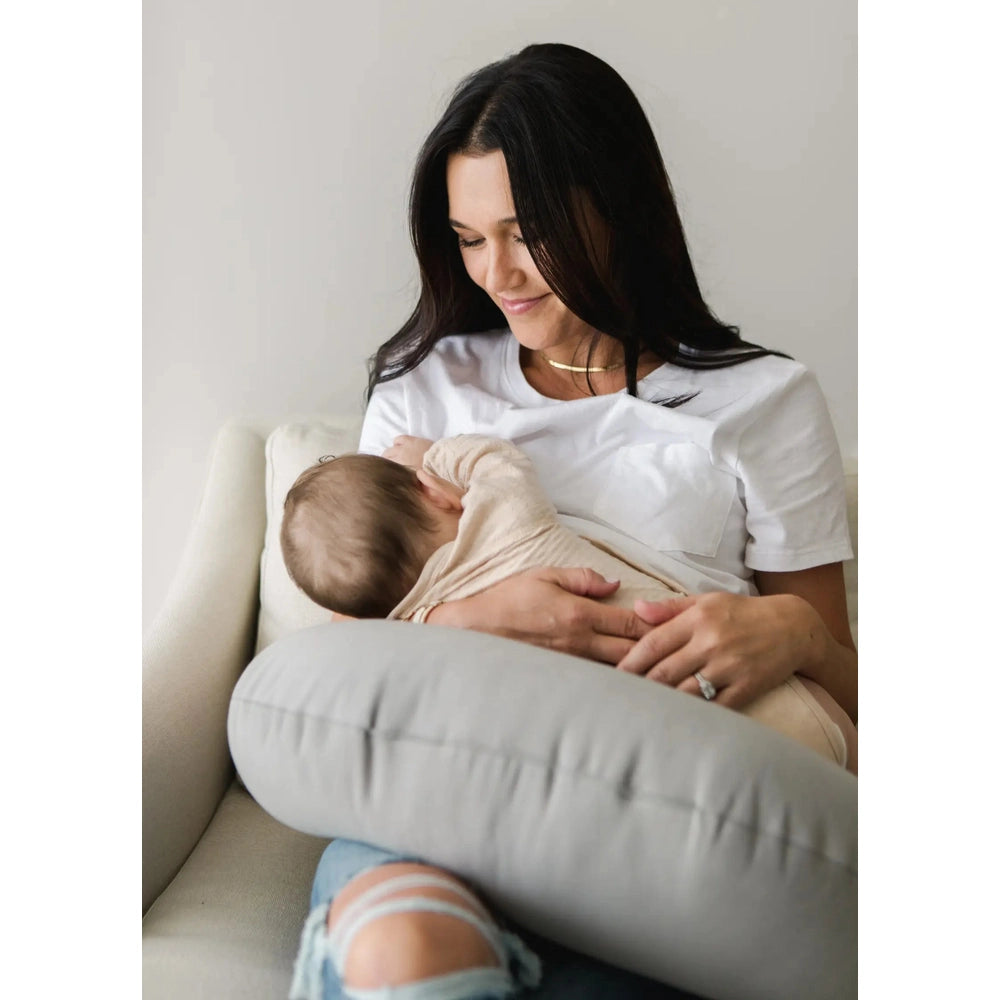 Snuggle Me Organic Feeding Support, cream nursing pillow, mother cradling baby on a sofa in a calm moment.
