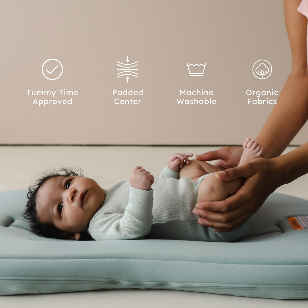 Snuggle Me Organic Lounger Curve in soft blue; baby lies calmly as caregiver supports legs, showcasing padded center and organic fabrics.