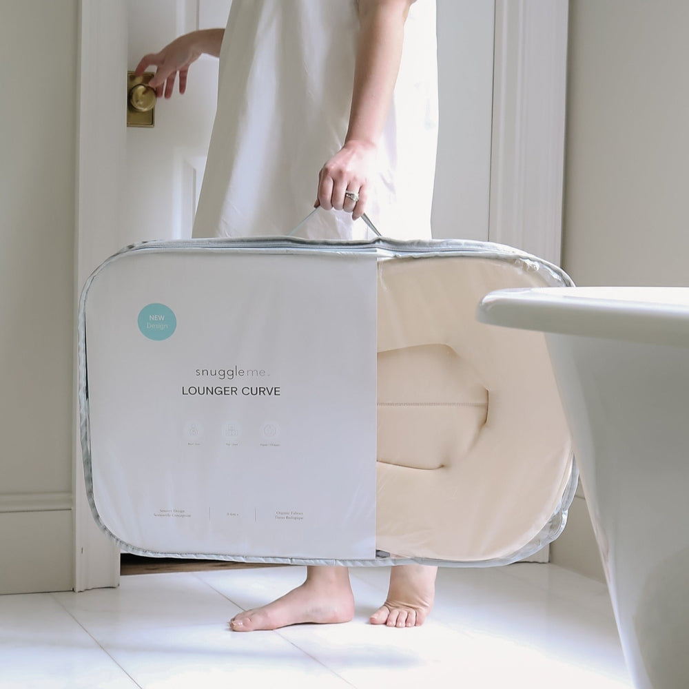 Person carries SNUGGLE ME ORGANIC Lounger Curve in light grey packaging through a bright doorway.