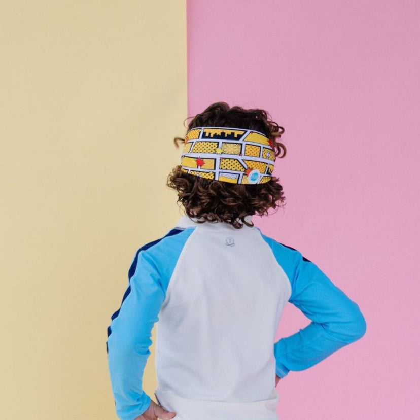 Splash Swim Goggles Comics child wearing blue and white swimwear with a comic-pattern headband against a pastel backdrop.