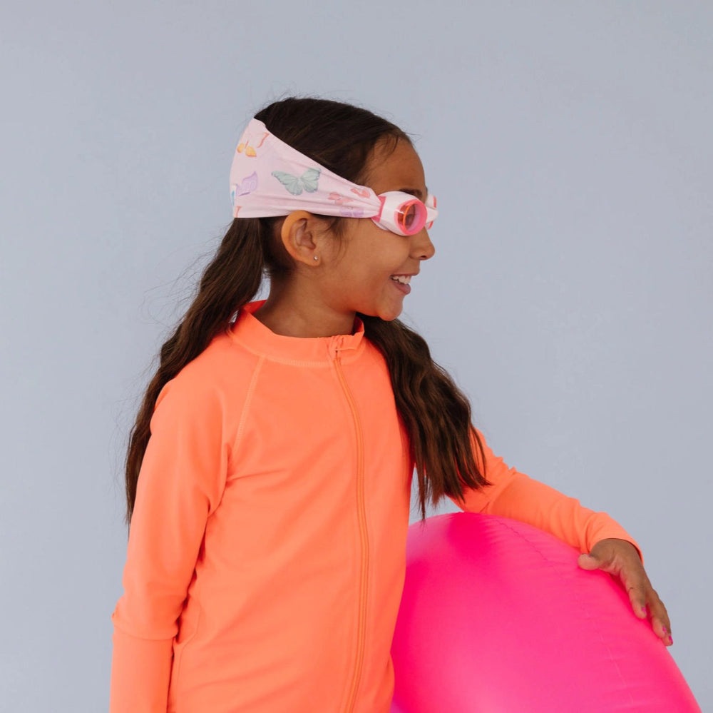 Splash Swim Goggles Flutter for kids; pink butterfly design goggles worn by a girl in a neon orange swimsuit with a pink float