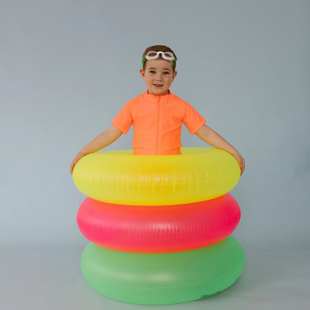 Splash Level Up Swim Goggles in White frames with green strap on a child in an orange rash guard, beside neon yellow, pink and green inflatables.