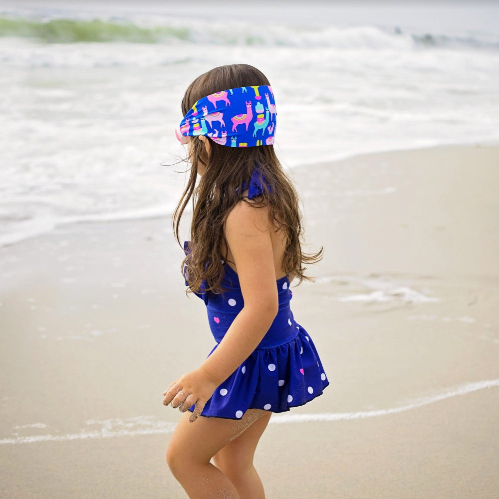 Splash Swim Goggles Llama Fiesta worn by a child on the beach in a blue polka-dot swimsuit and colorful llama headband