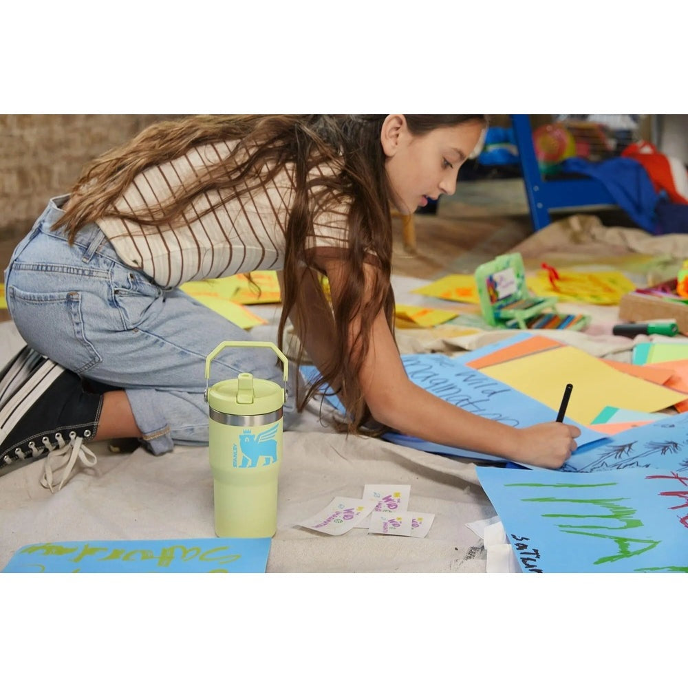 Stanley The Wild Imagination Iceflow Flip Straw Tumbler 20 oz in light green with carry handle sits on art-filled floor as girl colors posters