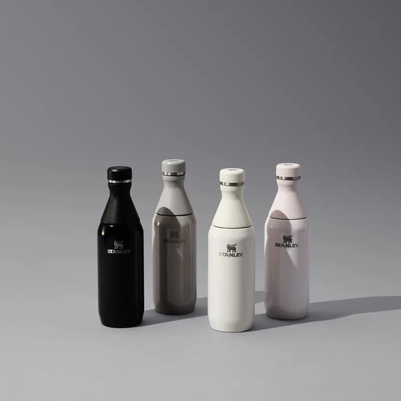 Four small decorative bottles with narrow necks and stopper tops, shown on a gray studio background. The bottles are black, gray, white, and pale pink, each with a small metallic logo on the front.