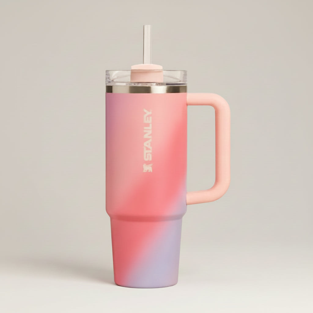 Stanley insulated travel tumbler with a handle, clear lid, and straw. The tumbler features a pink ombre gradient transitioning from coral pink at the top to a soft lavender purple at the bottom, with a white Stanley logo printed vertically on the side.