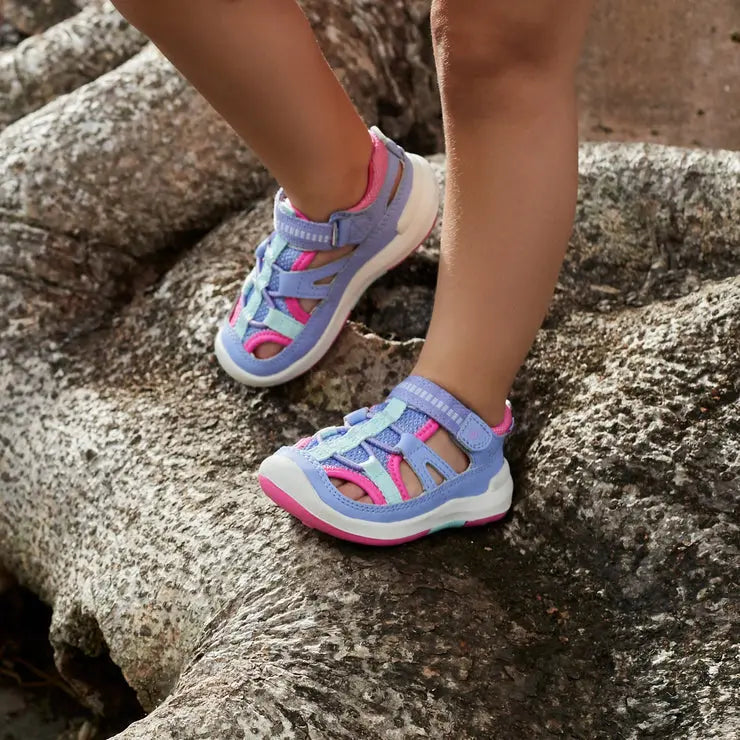 Stride Rite Strech Wade sneaker sandal for little kids in periwinkle with pink accents, strapped on rocky terrain.