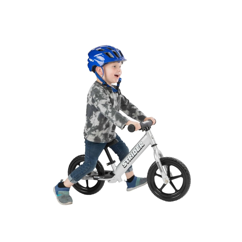 Strider 12 Pro Balance Bike, silver frame with black wheels, child balances and rides in a blue helmet.