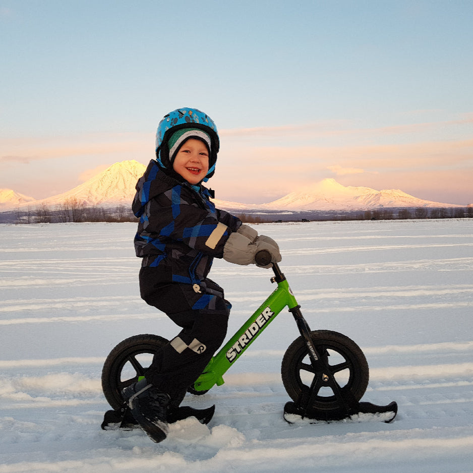 Strider 12 Snow Skis set; child riding a green Strider balance bike on snowy field in blue jacket and helmet