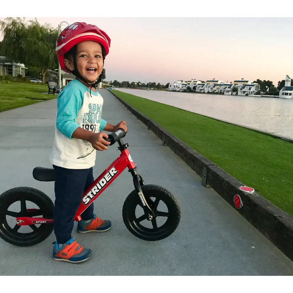 STRIDER 12 Sport balance bike, red frame with black wheels, being ridden by a smiling child in a red helmet by the lake.