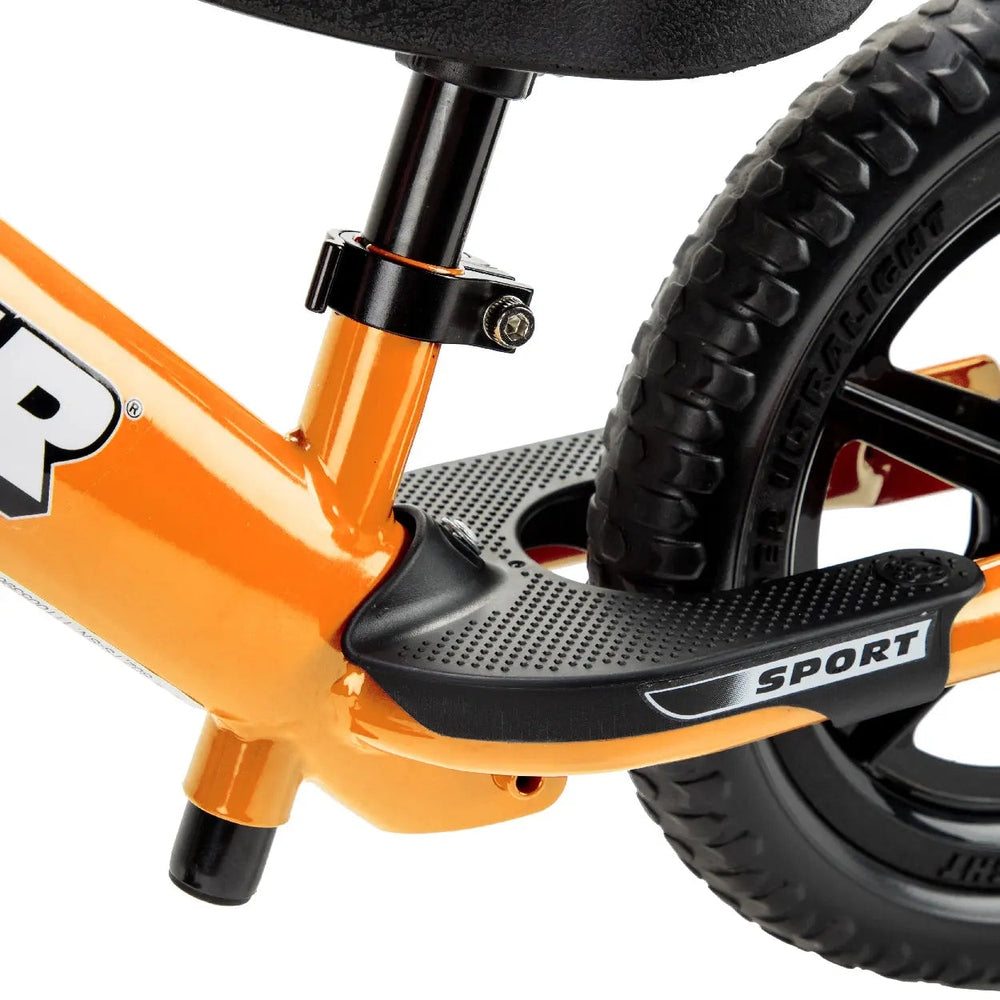 STRIDER 12 Sport balance bike, orange frame with a textured black SPORT footrest in use and chunky tires.