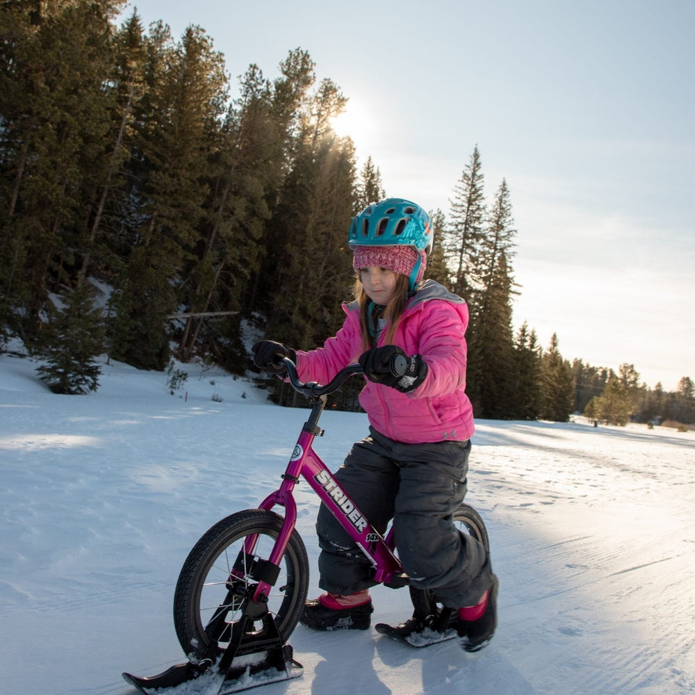 Strider 14X Sport Snow Skis set; pink frame, child in pink jacket riding on snowy trail with blue helmet.