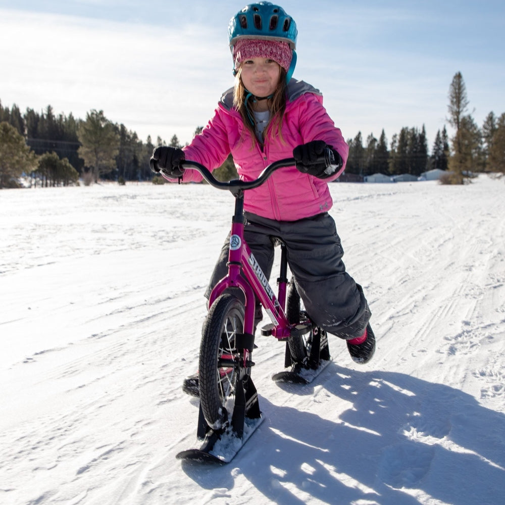 Strider 14X Sport Snow Skis set with pink frame; child rider in pink jacket and blue helmet on snowy field