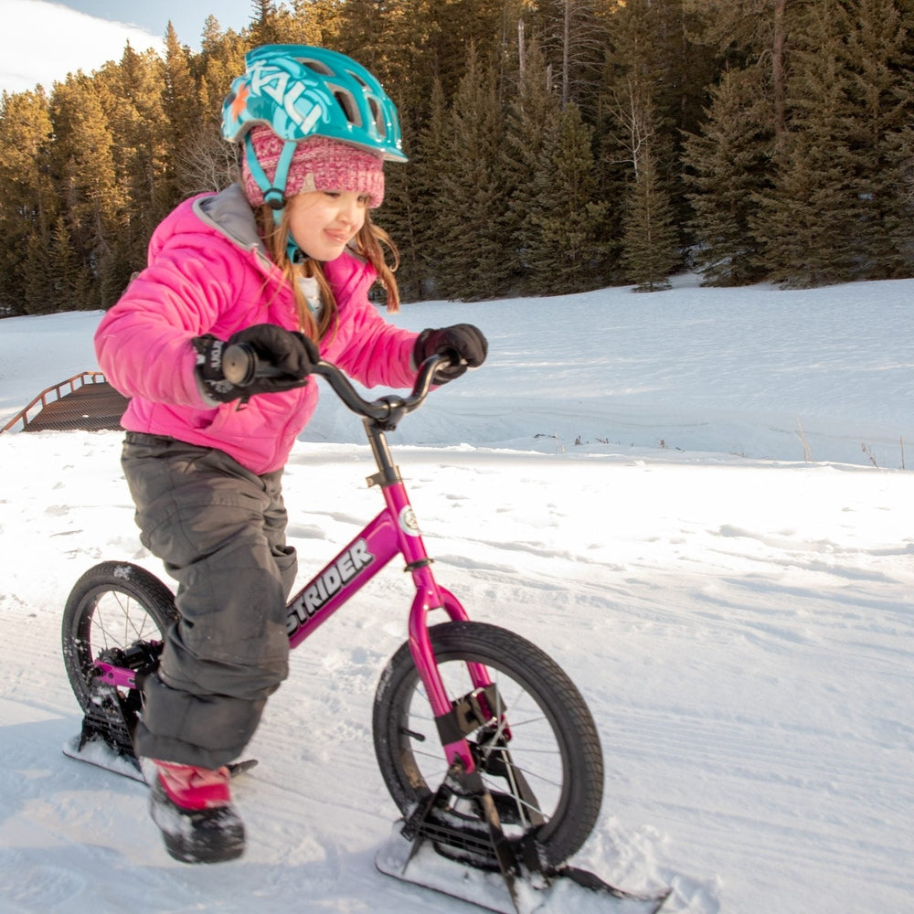 Strider 14X Sport Snow Skis set; pink frame, child in turquoise helmet riding on snowy ground