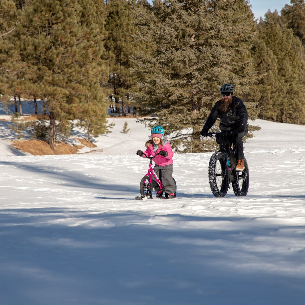 Strider 14X Sport Snow Skis set for kids, pink frame and blue helmet, outdoors with an adult cyclist in snow