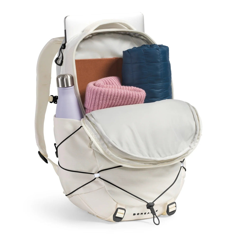 The North Face Borealis Backpack in Cream with pocket open, pink knit, brown notebook, blue sleeping pad, water bottle.