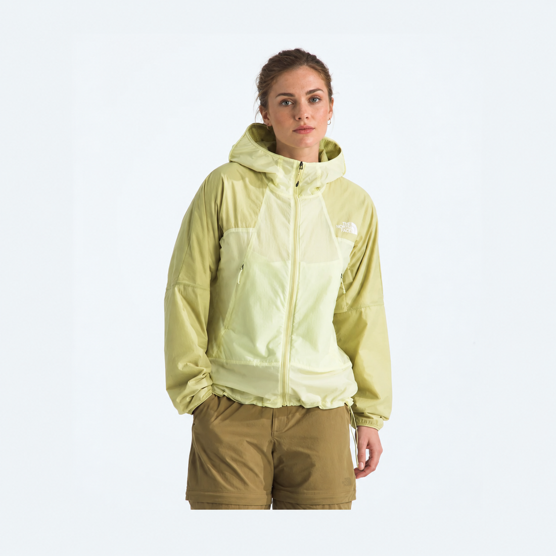 A woman modeling a light yellow-green hooded jacket with a white embroidered The North Face logo on the chest, paired with brown shorts, against a plain white studio background.