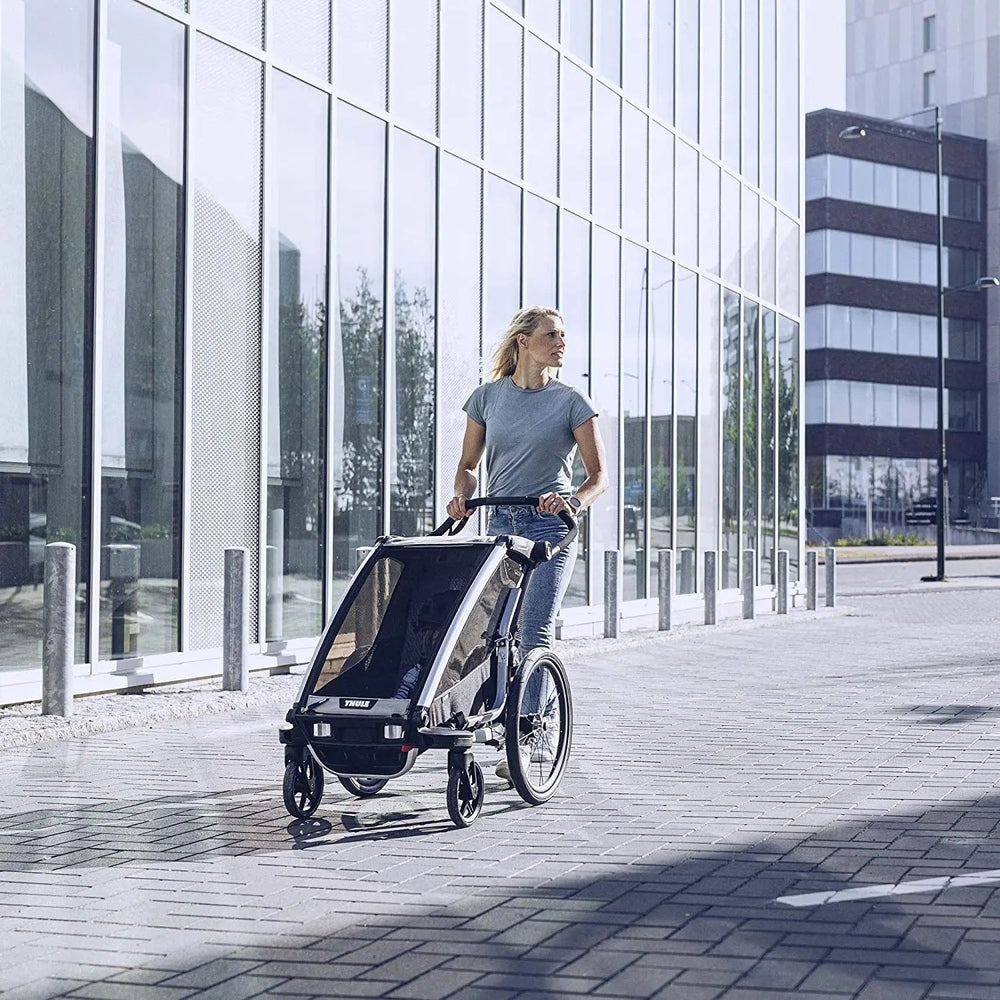 Thule Chariot Lite Multisport bike trailer Agave Green, being pushed on a city sidewalk by a woman.