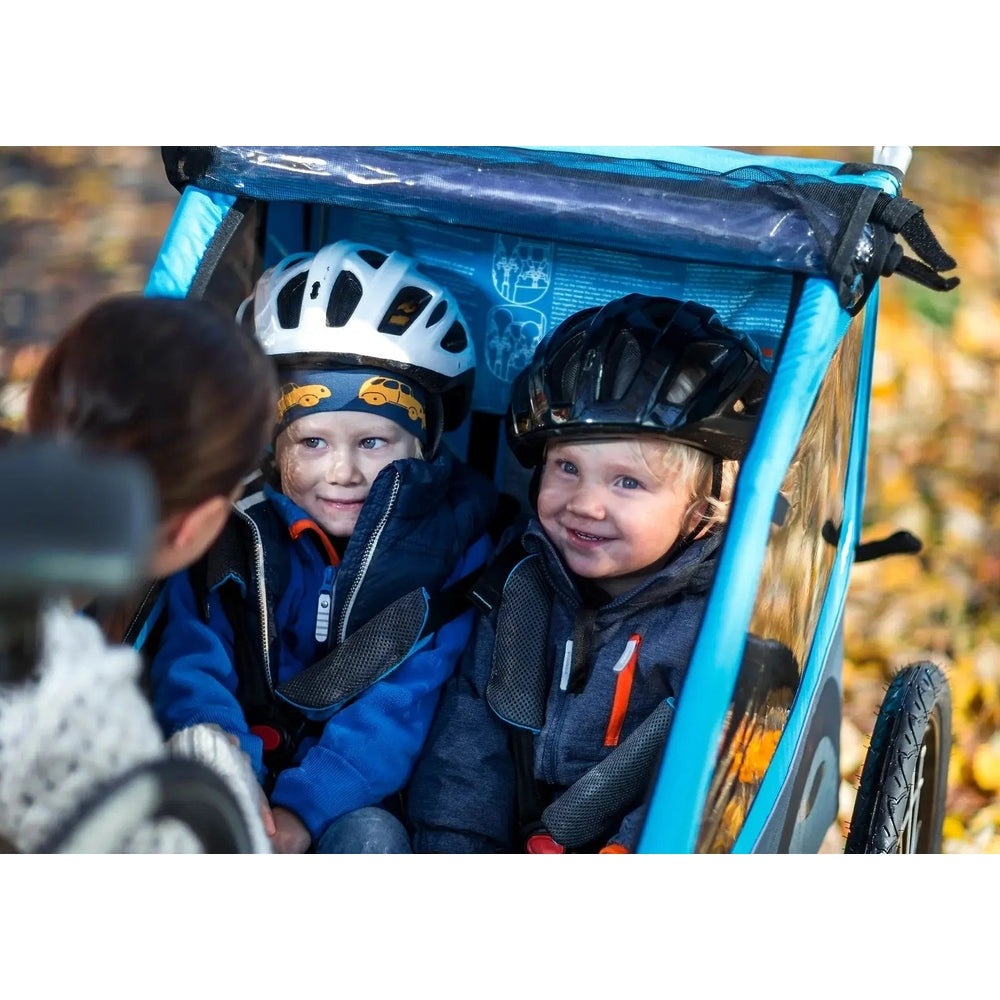 Thule Coaster Xt bike trailer for two kids; blue, being towed by a parent with smiling helmeted toddlers.