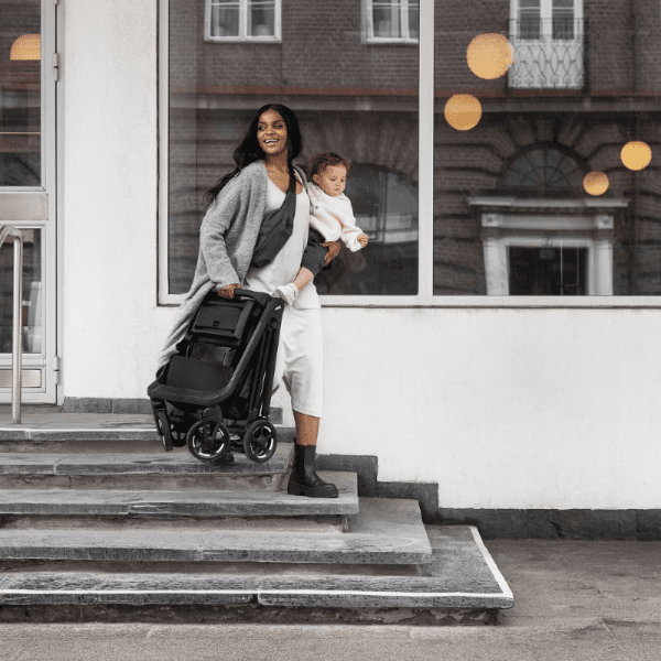 Thule Shine stroller in black folded frame carried by a smiling woman with baby on city steps