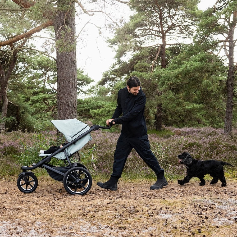 Thule Urban Glide 3 stroller in gray with black frame, being pushed by a man through a forest as a dog trails.