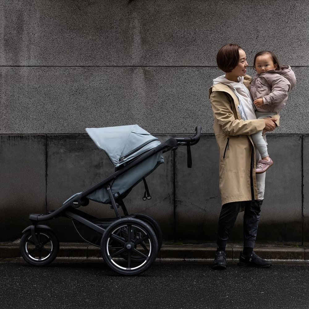 Thule Urban Glide 3 stroller in gray with canopy, beside a smiling mom carrying her toddler against a concrete wall.