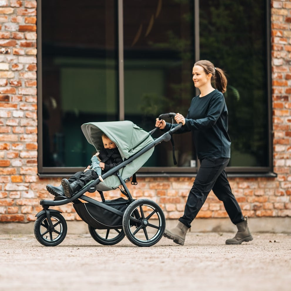 Thule Urban Glide 3 stroller in black with mint canopy, being pushed by a mother along a brick sidewalk.