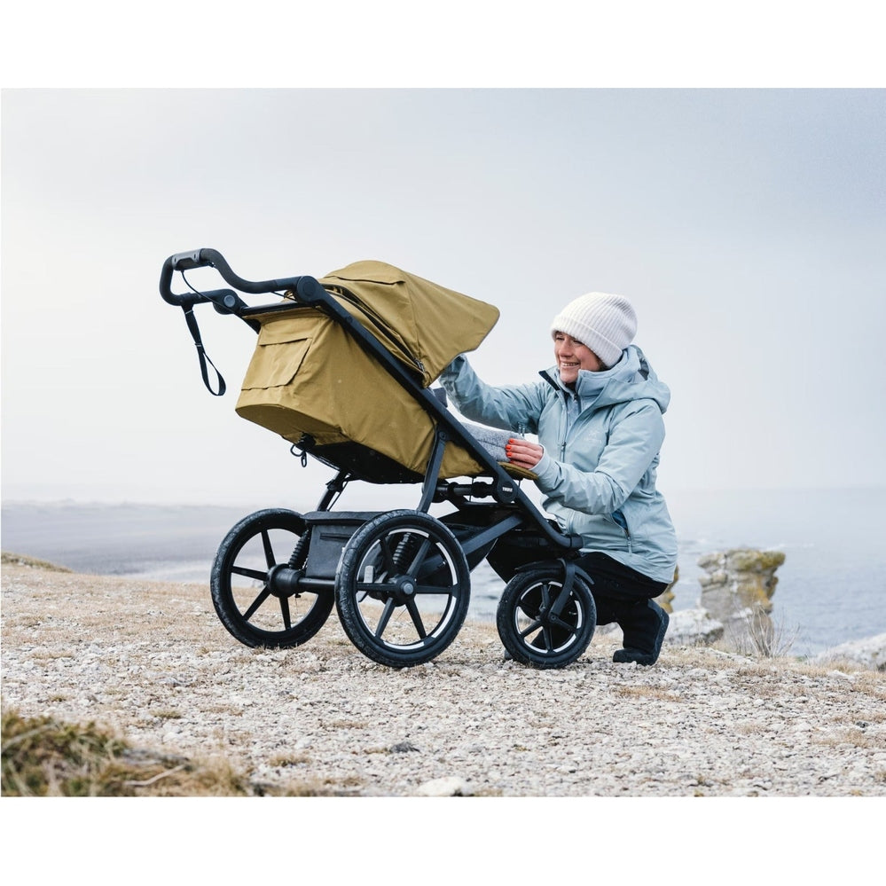 Thule Urban Glide 3 stroller with tan canopy on a black frame, pushed by a smiling person in a light blue jacket.