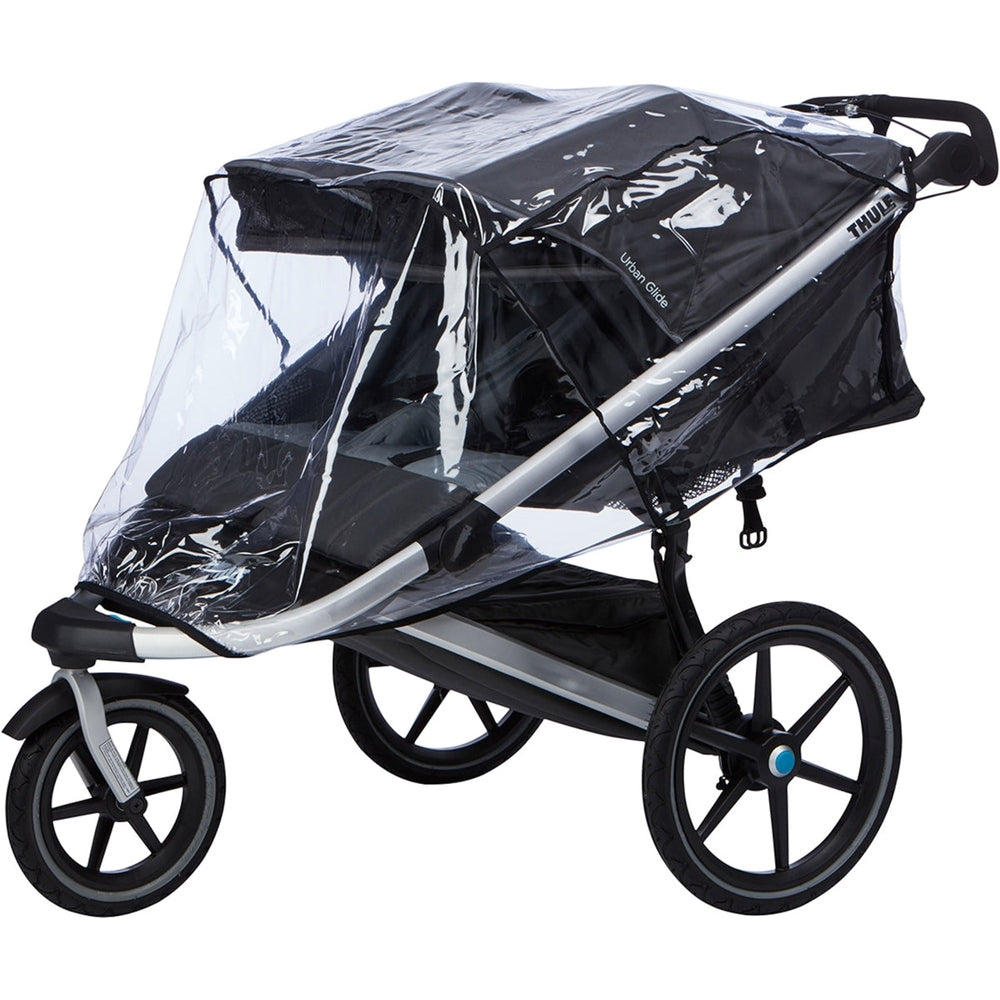 Thule Urban Glider Double Rain Cover shields a black stroller with a clear canopy while a parent pushes.