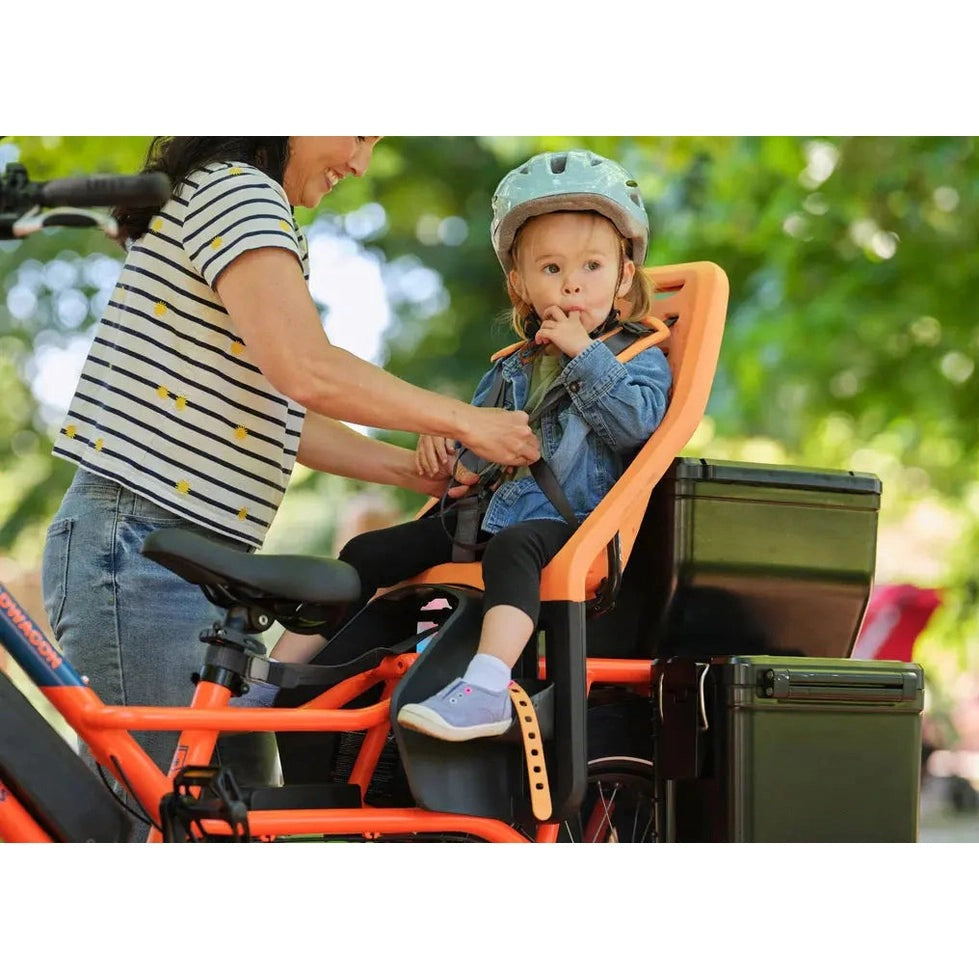 Thule Yepp Maxi rack-mounted child bike seat in orange, with toddler strapped in and helmeted while a mom buckles the harness.