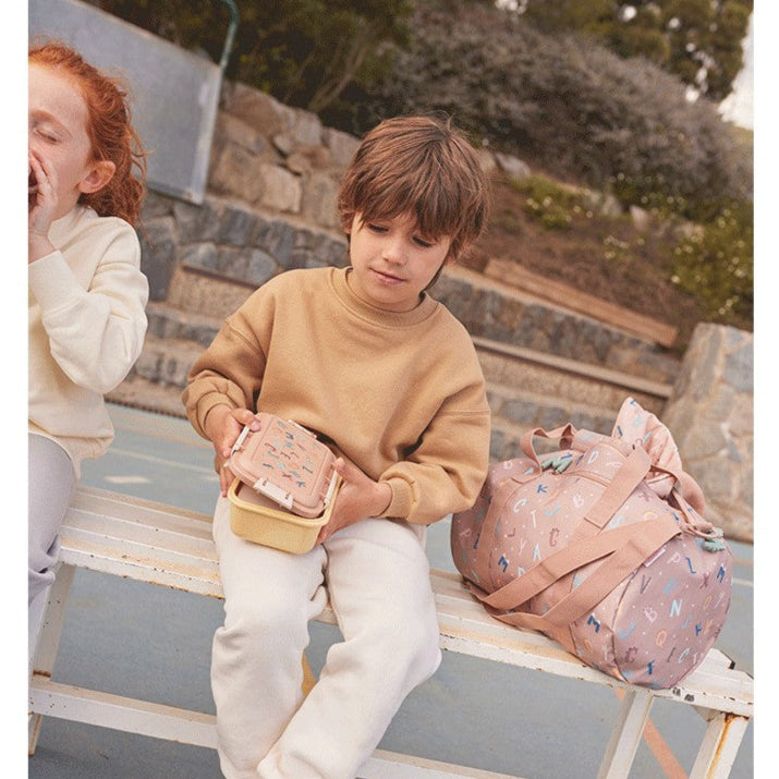 Tutete Kids Bento Lunch Box pink with pastel yellow inner tray, held by a child next to a pink backpack.