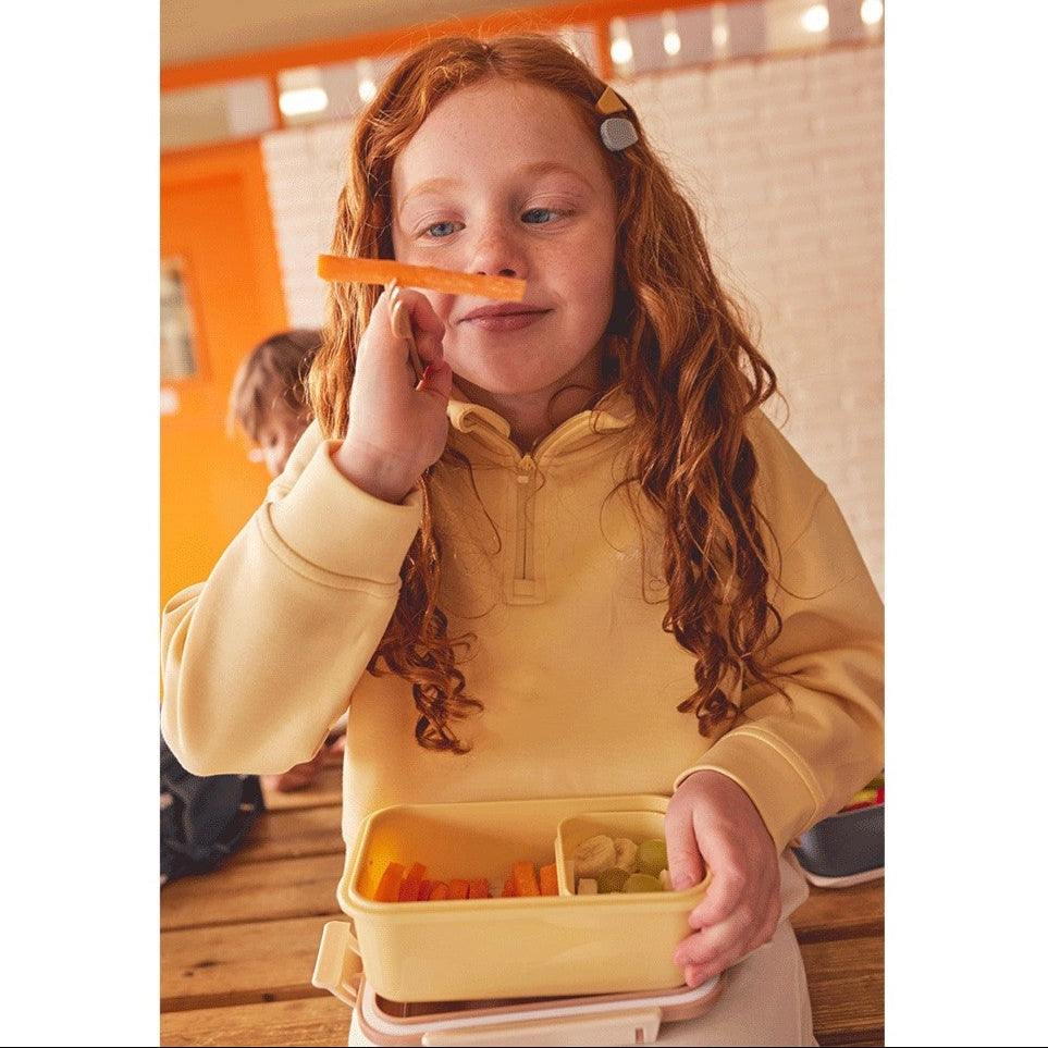 Tutete Kids Bento Lunch Box in pale yellow with divided compartments; girl holds carrot stick.