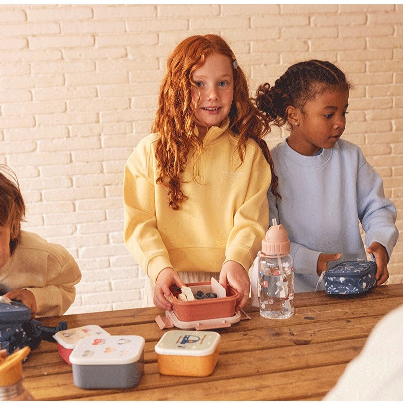 Tutete Kids Tritan Water Bottle 500 ml with pink lid, beside colorful lunch boxes on a wooden table