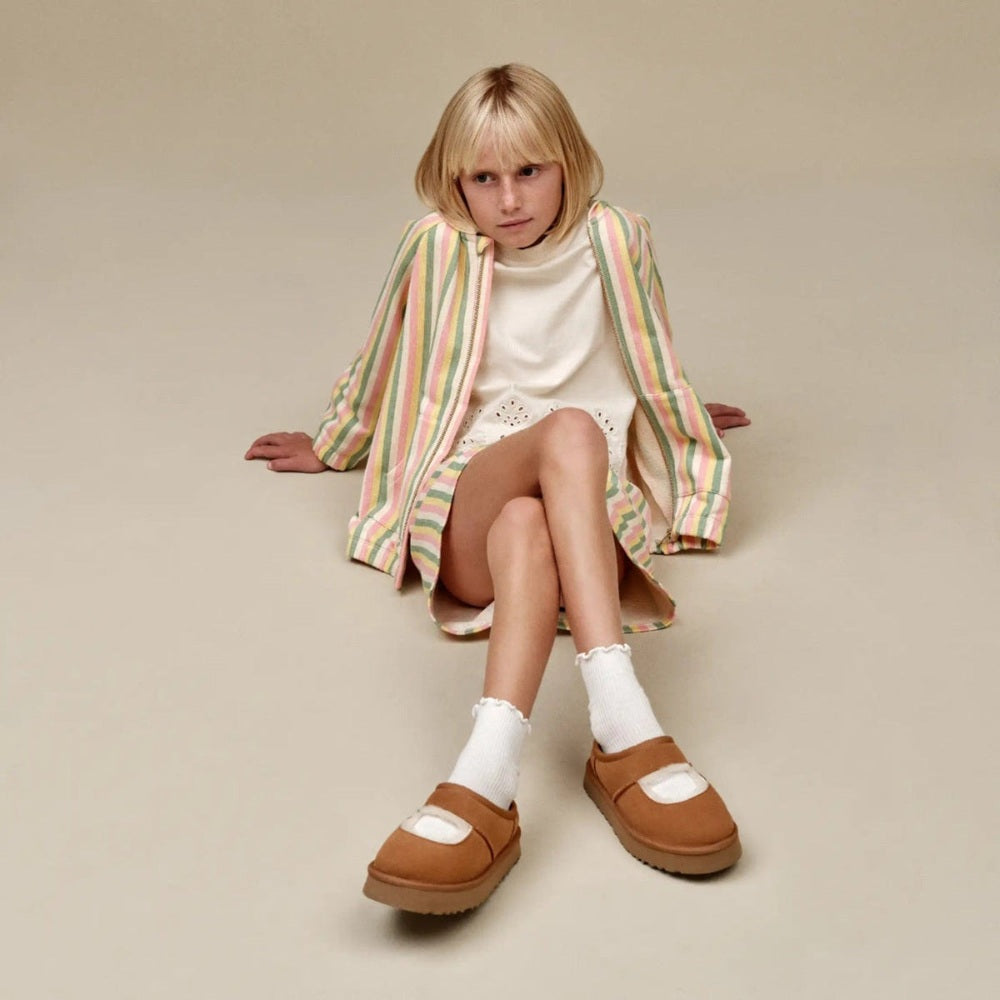 UGG Kids' Bea Mary Jane Chestnut shoes worn by a seated girl in a pastel striped coat and white dress.