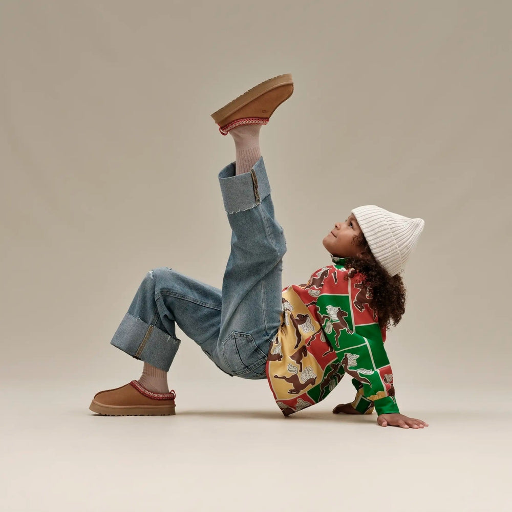 UGG Kids' Tazz Chestnut suede shoes; playful kid in denim lifts leg to show the flexible tan sole.