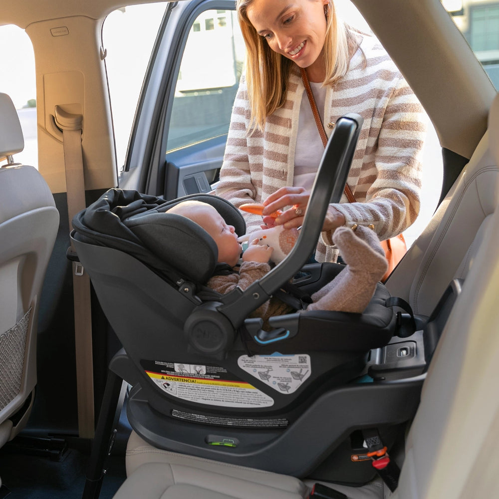 UPPAbaby Aria Car Seat; black/gray infant seat installed rear-facing in car as caregiver feeds baby.