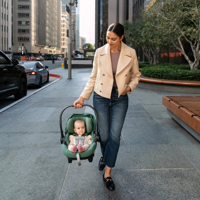 Uppababy Aria infant car seat in green, baby buckled securely, being carried by a mom on a city sidewalk.