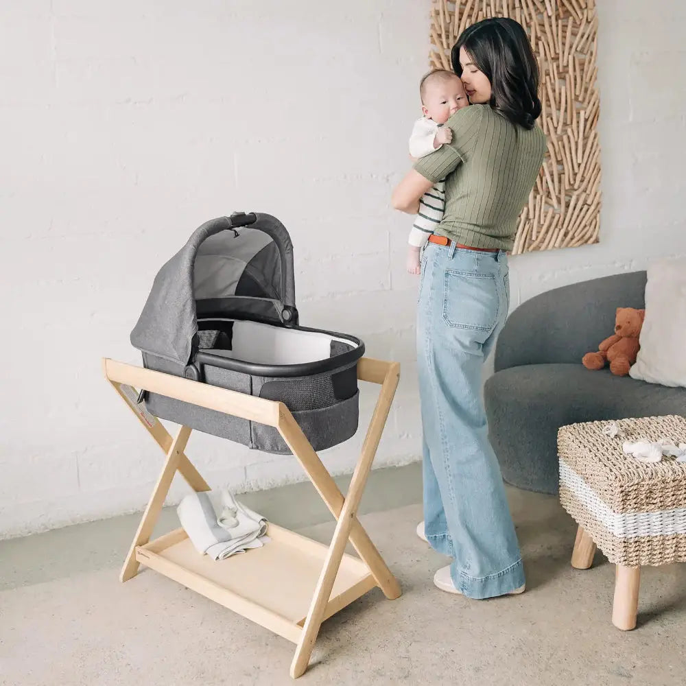 Uppababy Bassinet Stand with grey bassinet on a natural wood frame; mother cradles baby nearby.