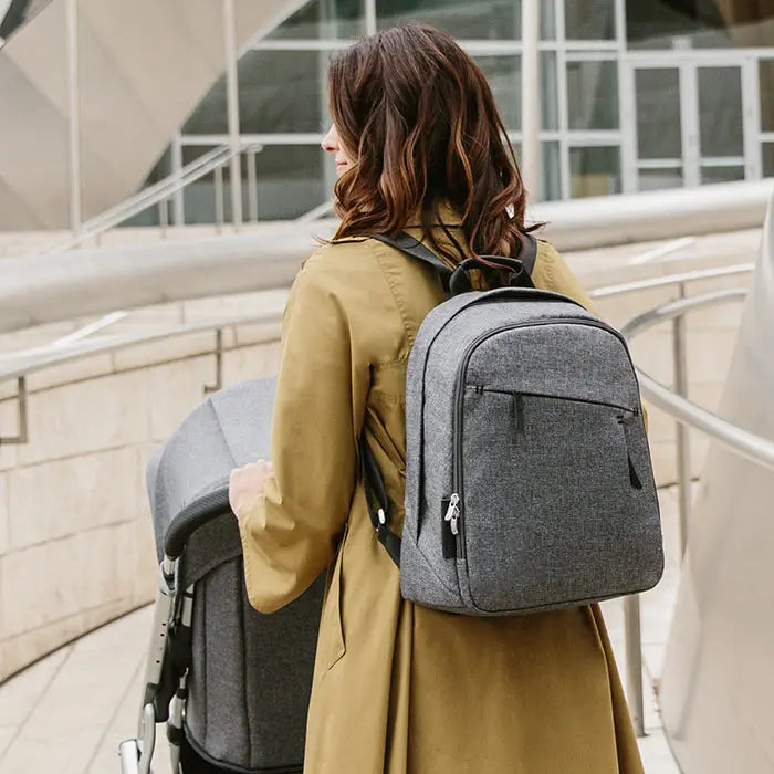 Uppababy Changing Backpack in gray fabric worn by a woman in a tan coat, showing front zip pockets.