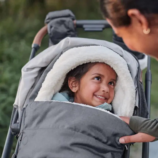 Uppababy Cozy Ganoosh stroller cover in gray with plush white lining, keeping a smiling baby warm as mom adjusts the cozy hood.