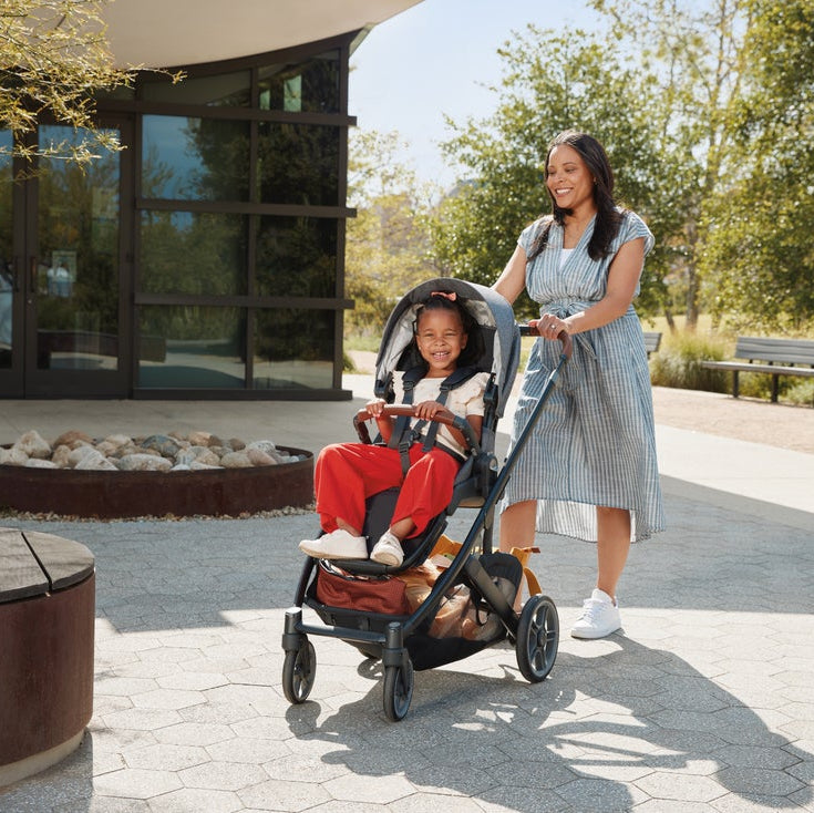 Uppababy Cruz V3 stroller with black frame and brown storage; mother pushing a smiling child in red pants.