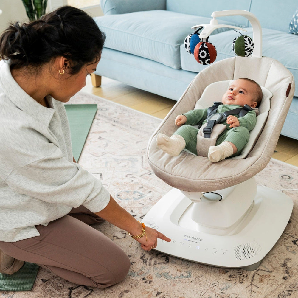 UPPABABY Mamaroo Smart Swing with beige fabric seat, baby secured in harness on white base with overhead toy mobile