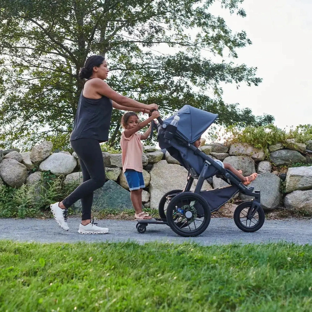 Uppababy Ridge Stroller in gray, mom pushing along a park path while a child stands on the footboard.