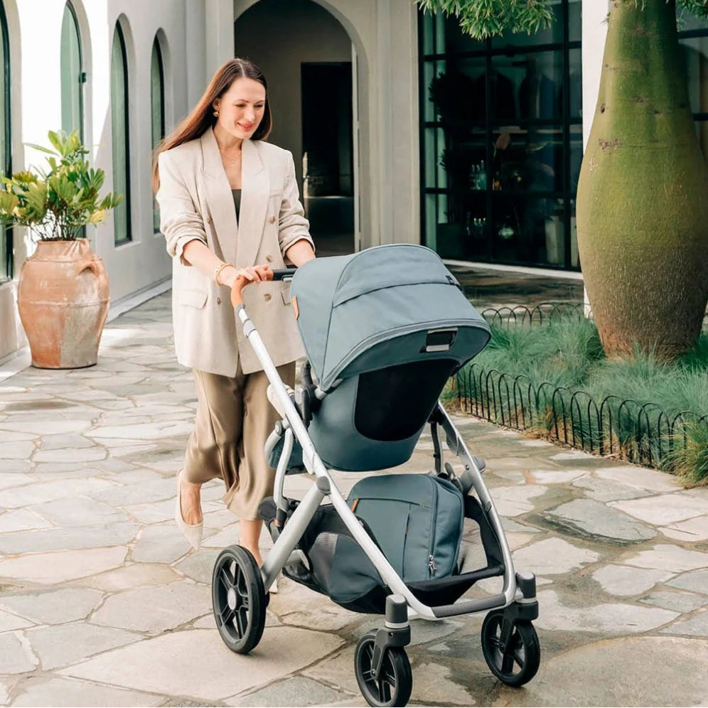 Woman pushing a stroller in an outdoor setting with a building and plants in the background