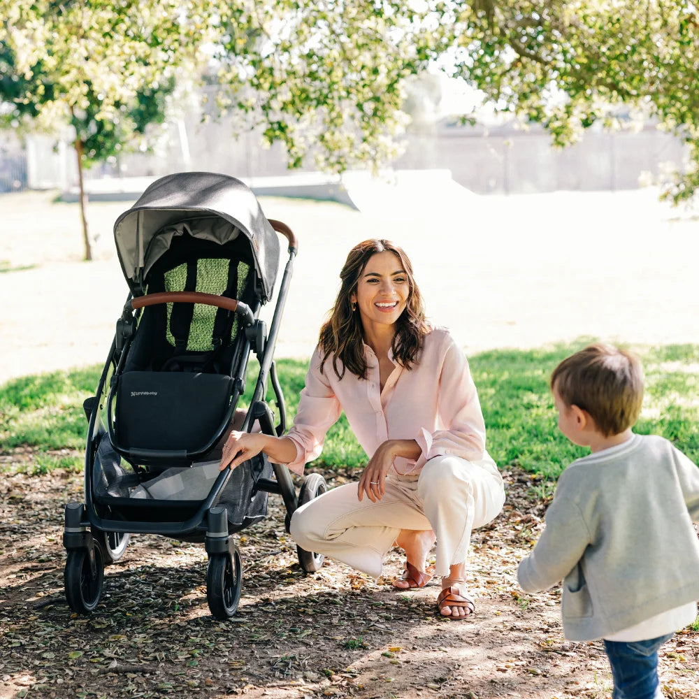 Uppababy Vista V3 Stroller in black with gray canopy and brown handle, mother kneels by it in a sunny park.