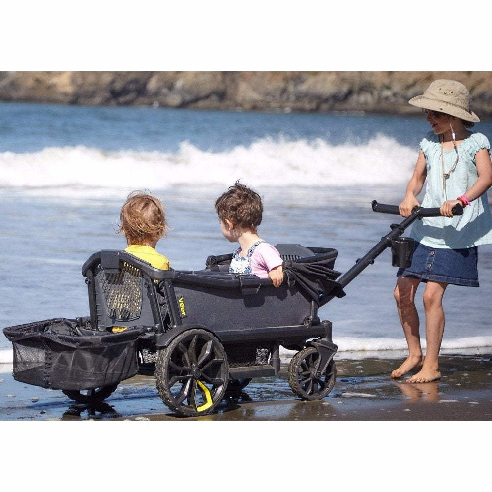Veer All-Terrain Cruiser Wagon, black ride-along with two kids seated, pushed by a woman on the beach.