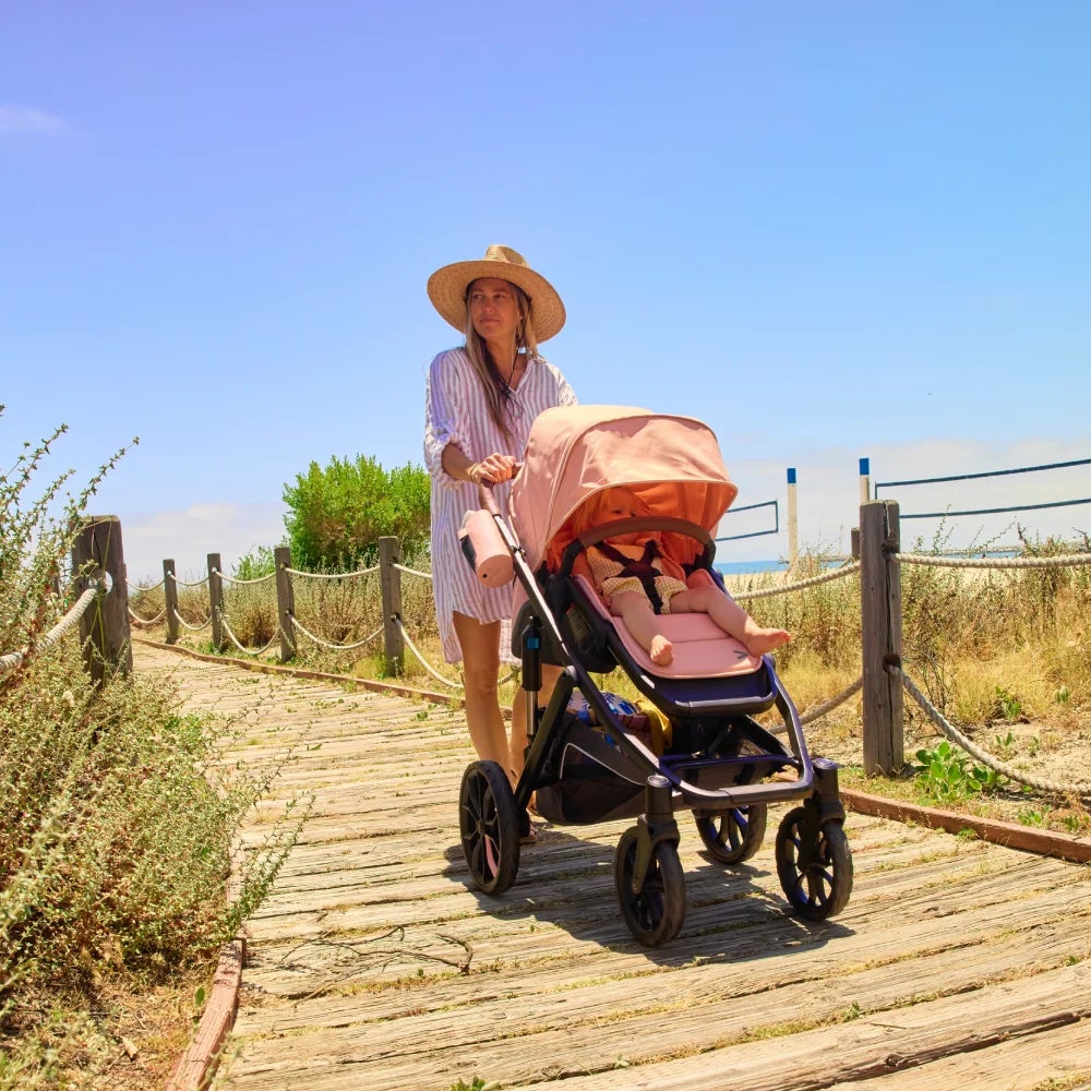 Woman pushing a pink stroller on a wooden path with a clear blue sky.