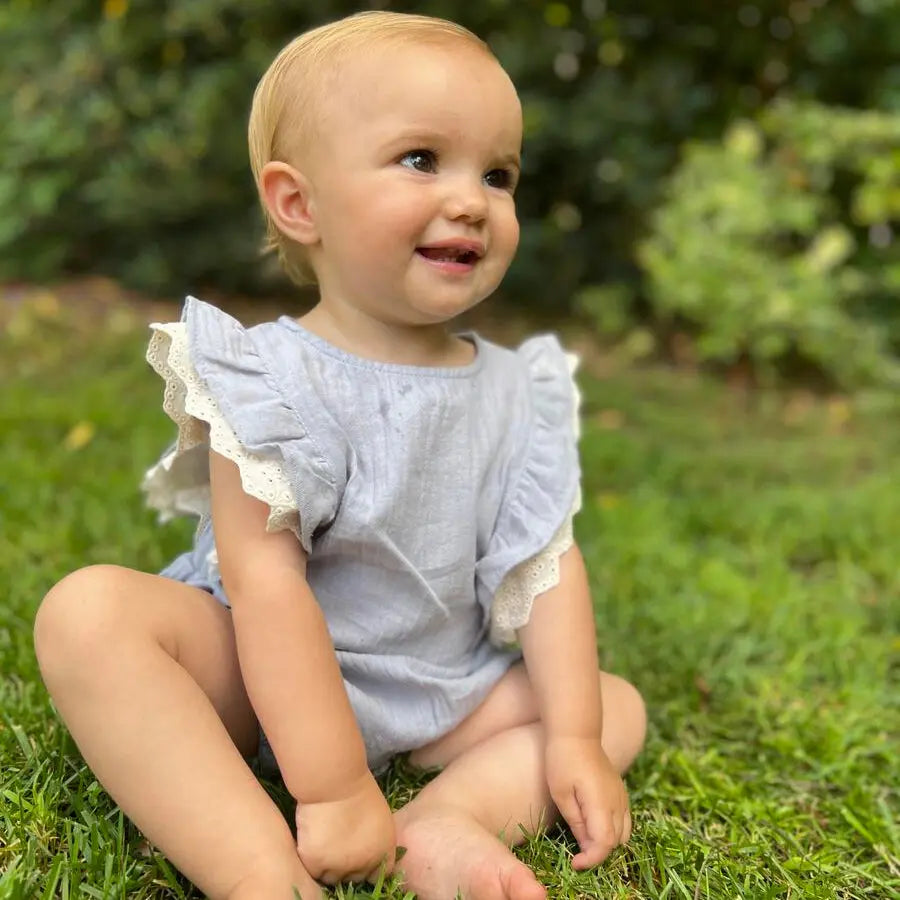 Vignette Megan Bubble Blue baby romper, light blue cotton with lace sleeves, worn by a smiling toddler on grass.